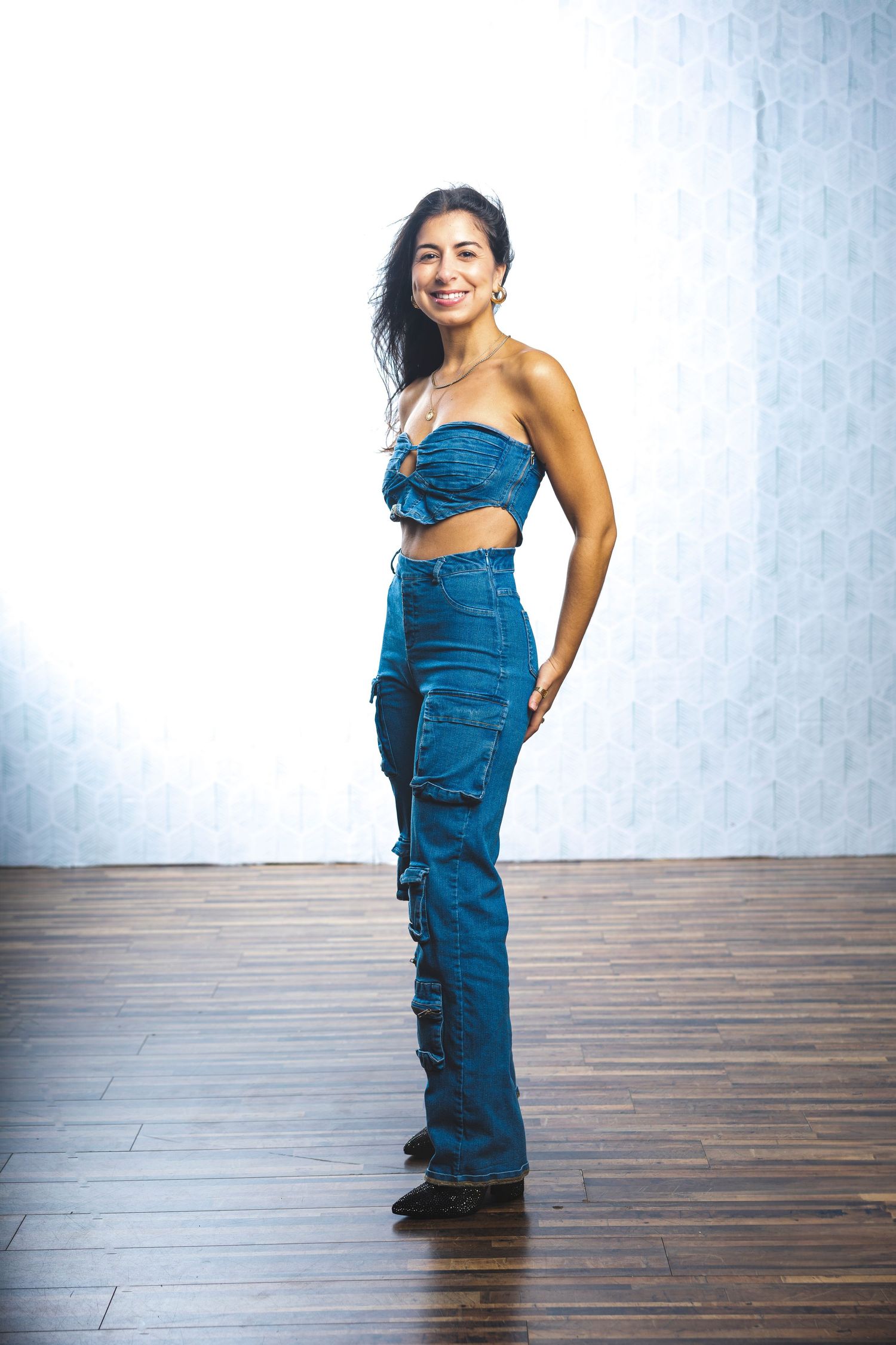 Model in blue denim outfit strikes confident pose on wooden floor against white wall.