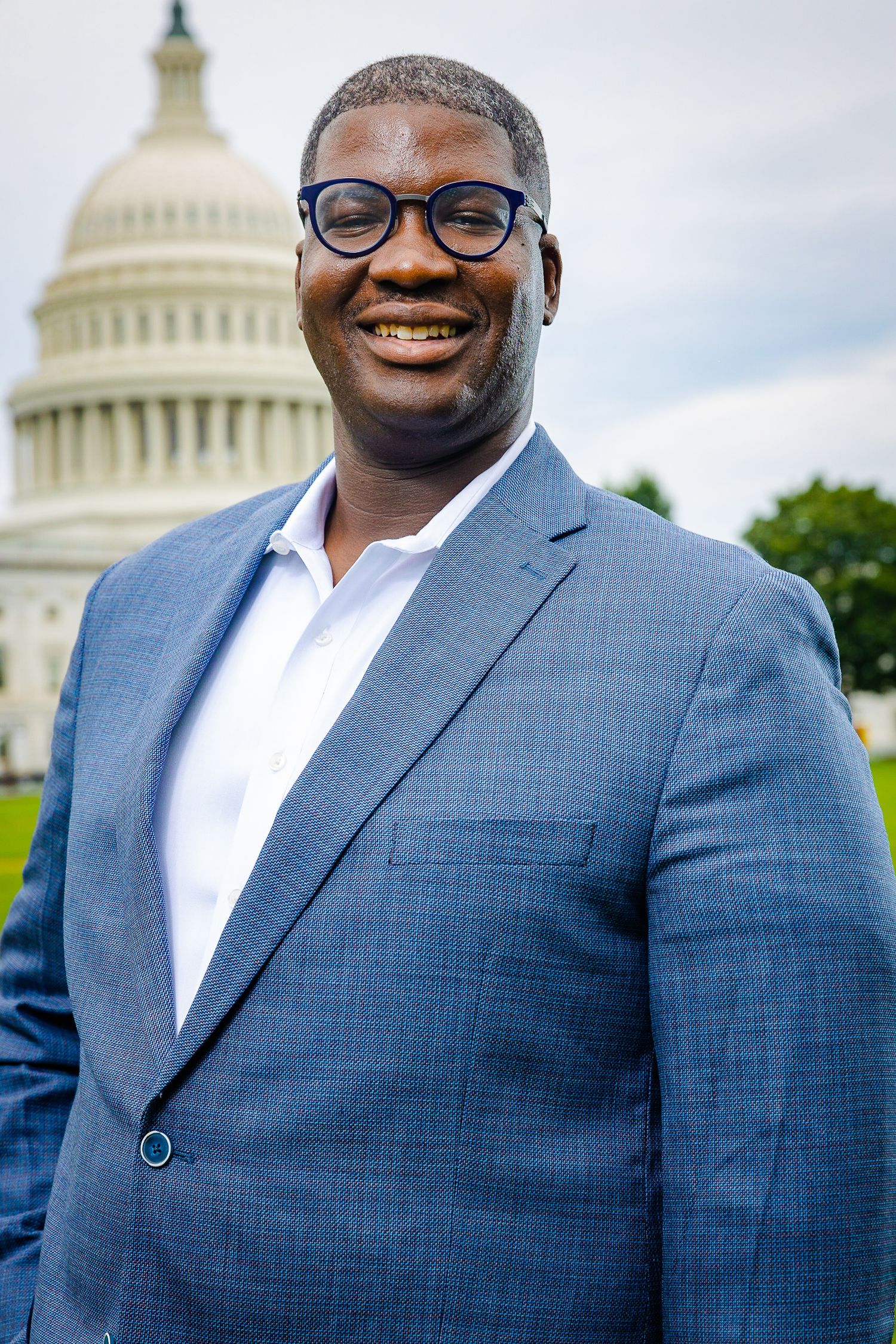A person wearing glasses and a blue suit jacket stands smiling in front of the US Capitol Building dome on a sunny day.