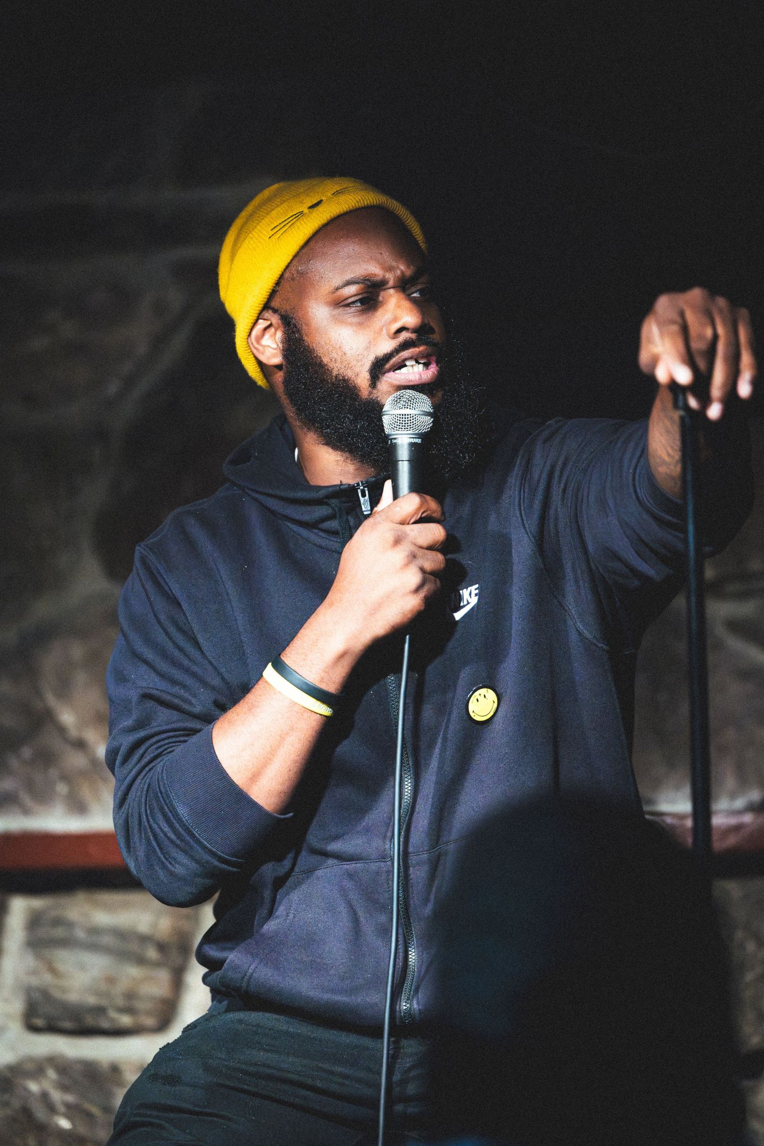 Performer in yellow beanie and navy hoodie speaks passionately into microphone during intimate standup comedy show.