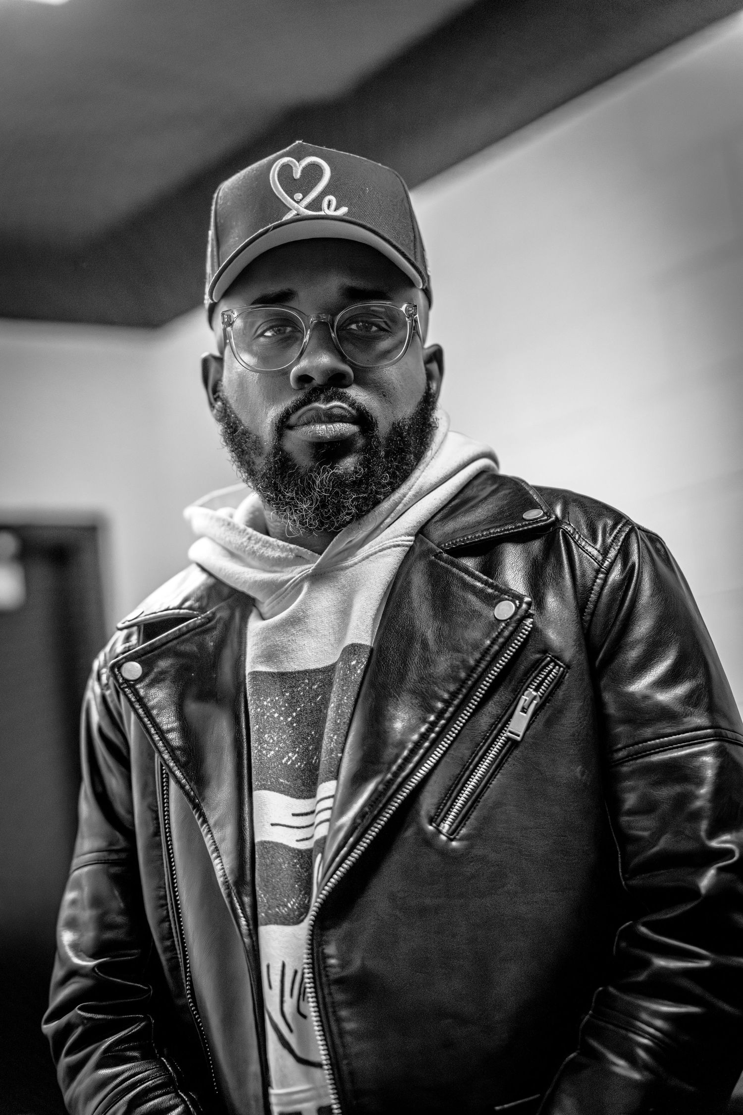 Black and white portrait of someone wearing a baseball cap, glasses, and leather jacket with white hoodie underneath.