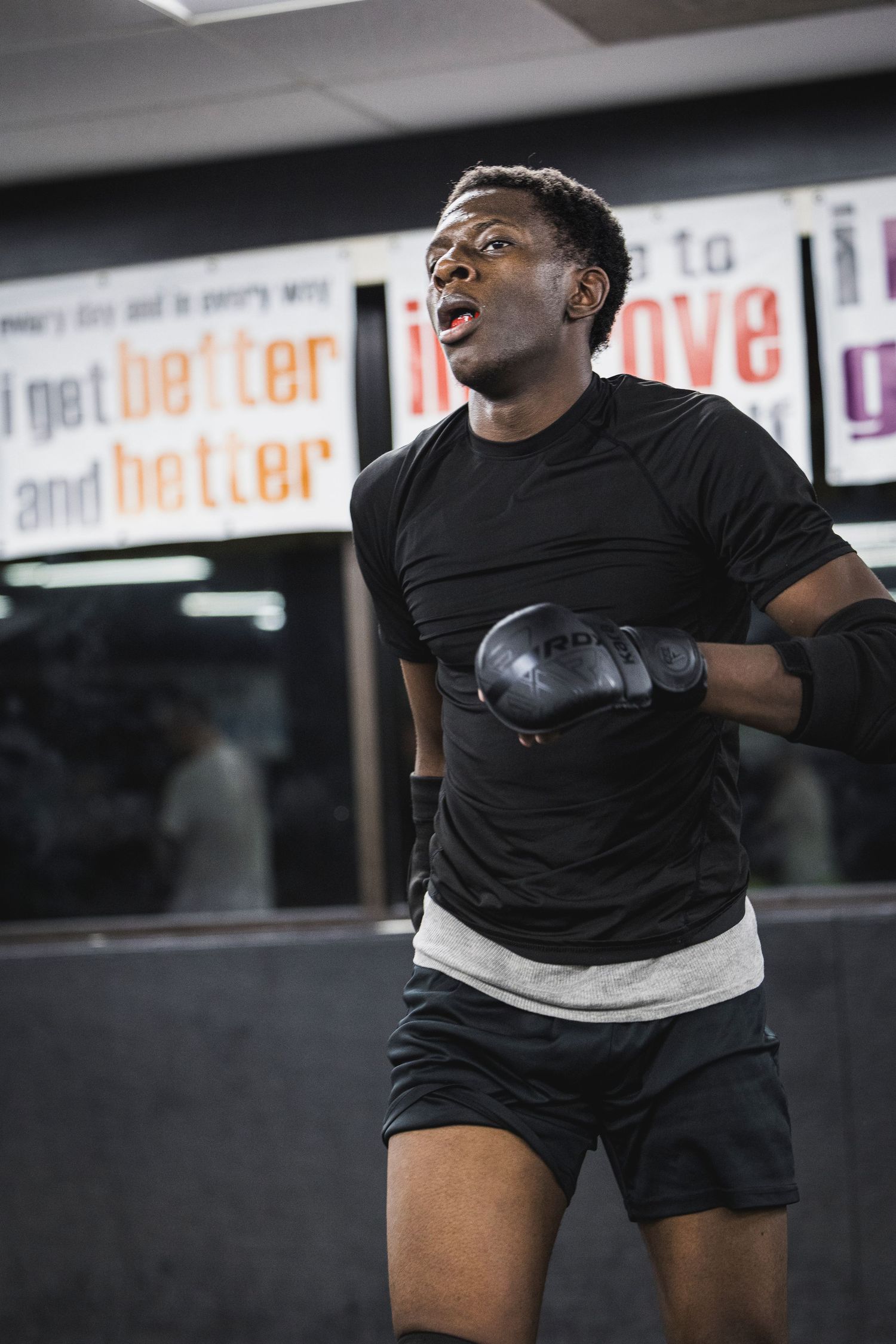 Athlete in black t-shirt and shorts wearing boxing gloves trains intensely at a fitness facility with motivational signs.
