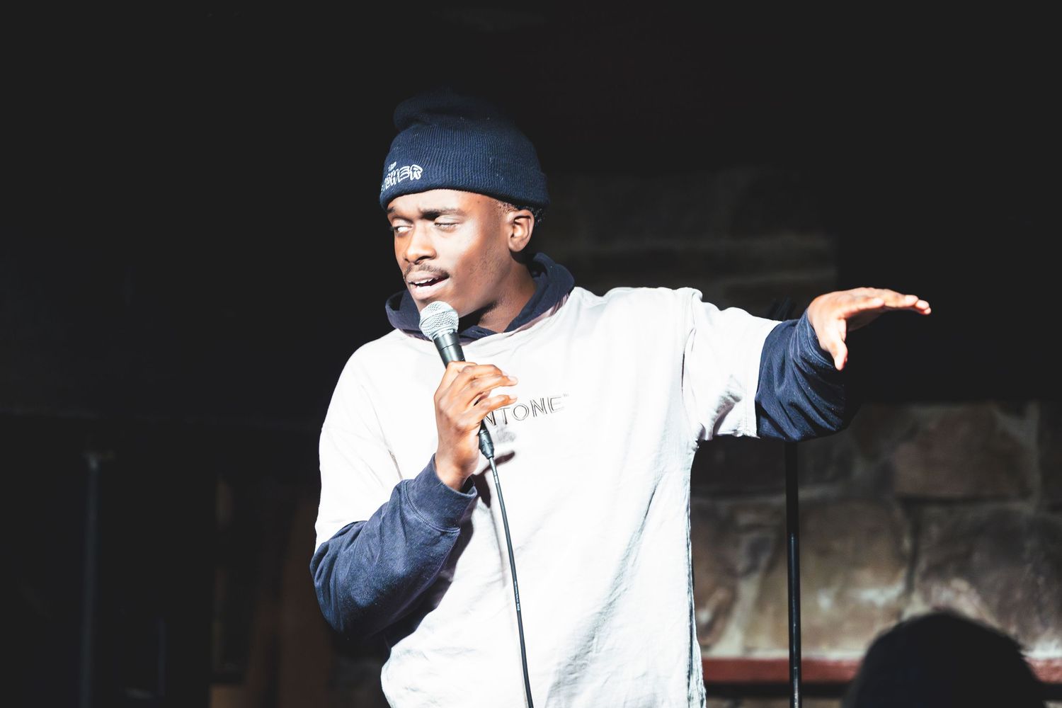 A performer in a white sweatshirt and navy cap holds a microphone while gesturing during a stand-up comedy show.