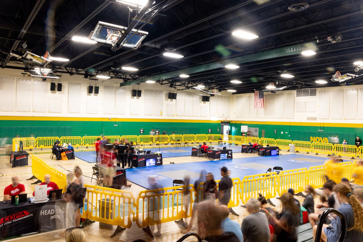 Indoor sports arena with blue wrestling mats surrounded by spectator seating in a gymnasium setting.