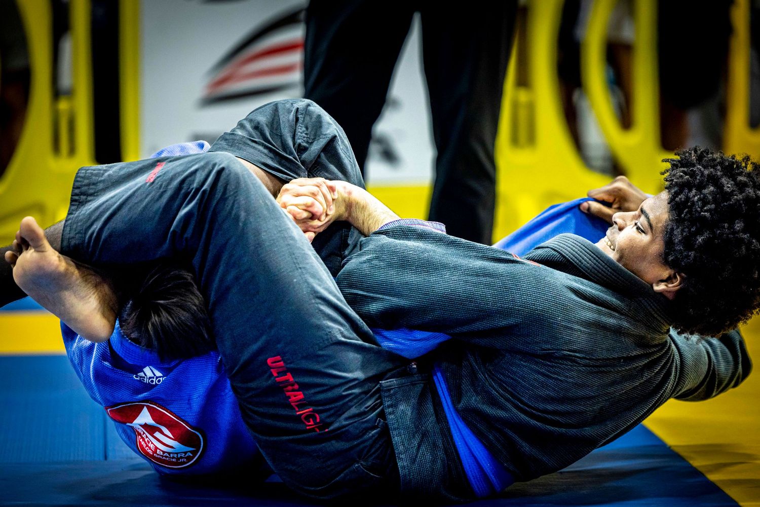 Two competitors in blue gis grappling on yellow-bordered competition mat.