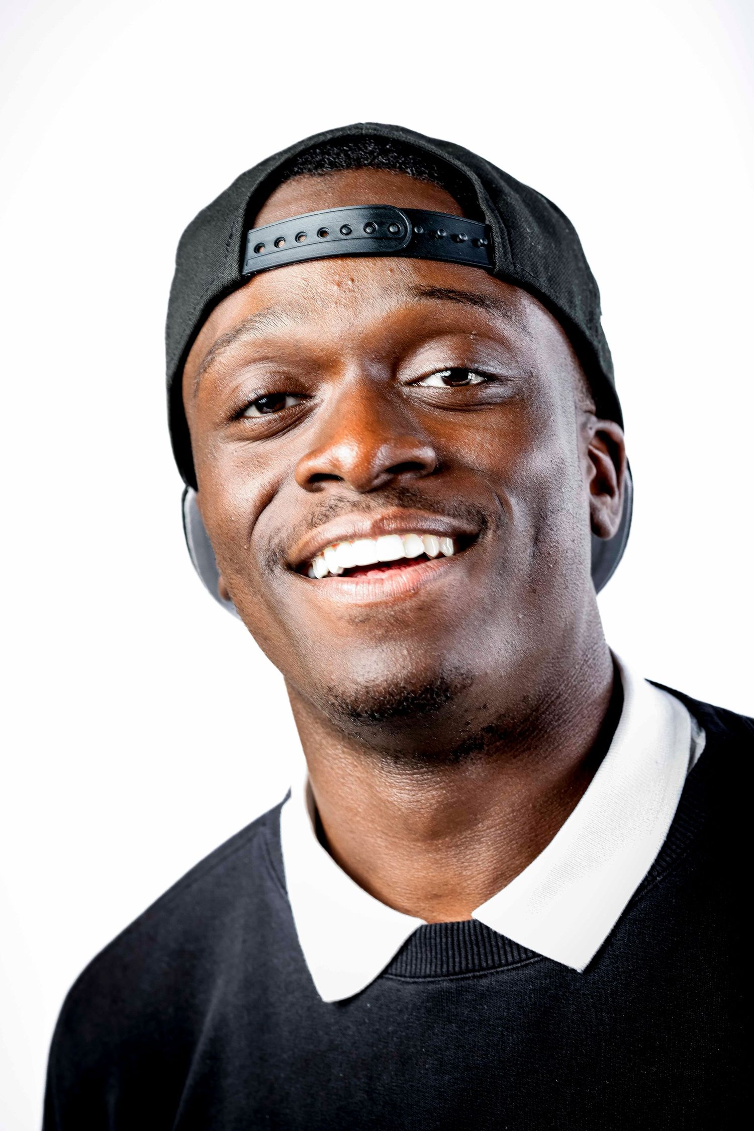 Person wearing black cap and white collar smiling brightly against white background.