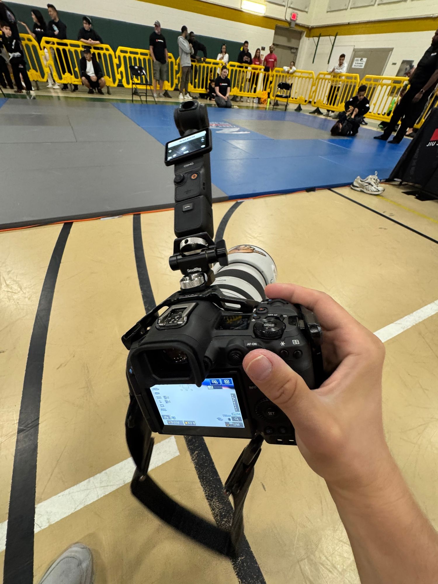 A DSLR camera with mounted flash is held at a gym with yellow barriers and blue mats visible in the background.