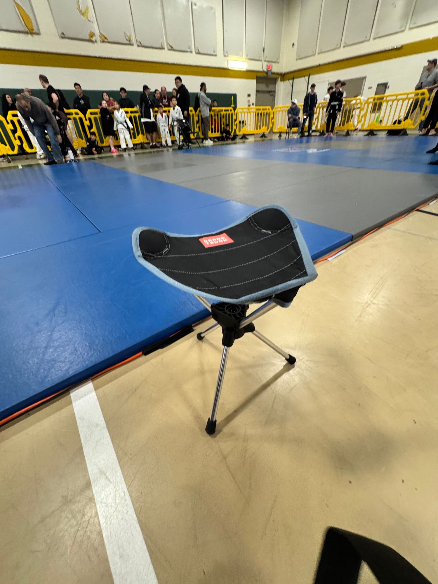 Portable black camping stool sits on blue wrestling mat in gymnasium with yellow railings visible in background.