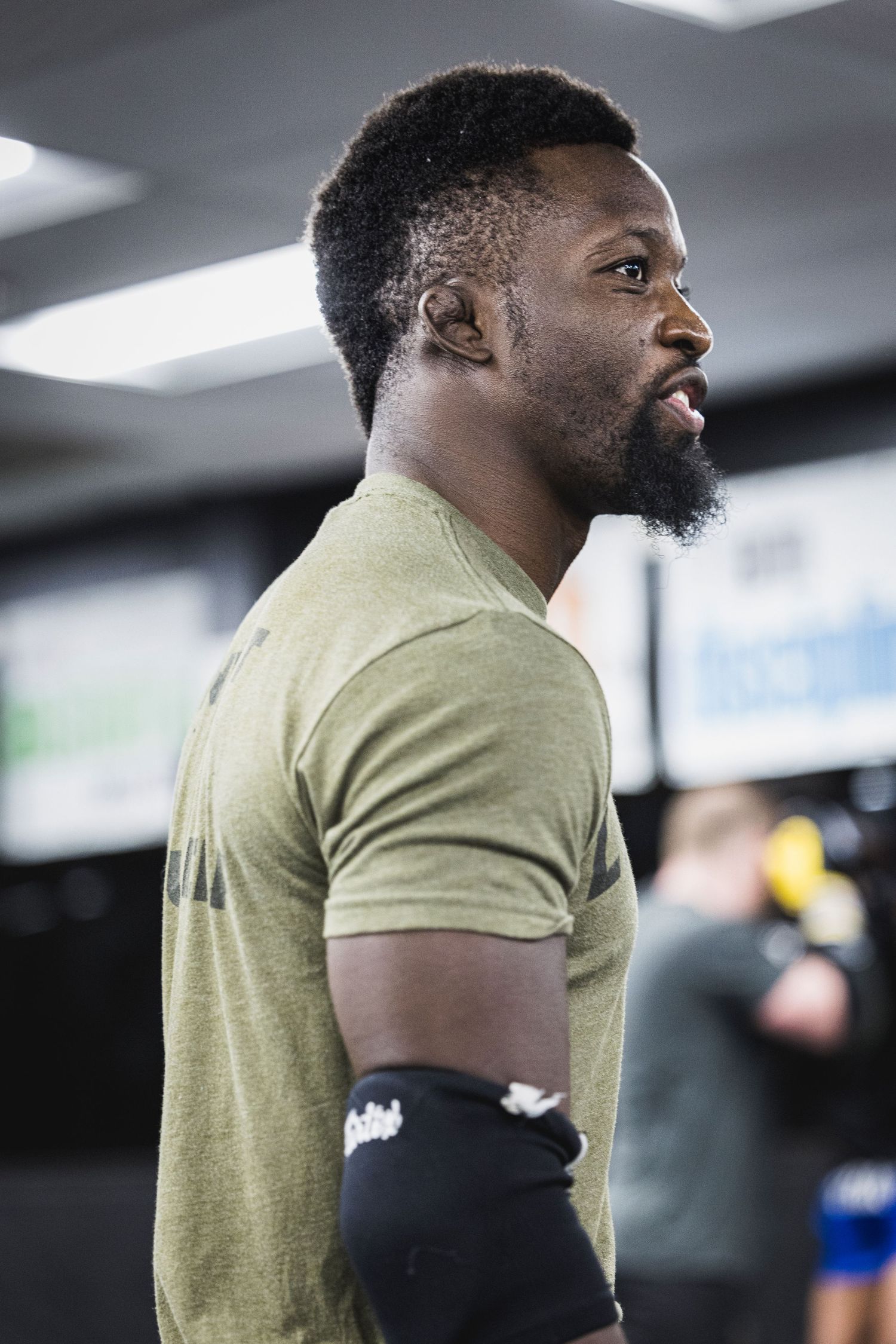 Athlete in an olive green t-shirt and arm sleeve stands in profile at a training facility.