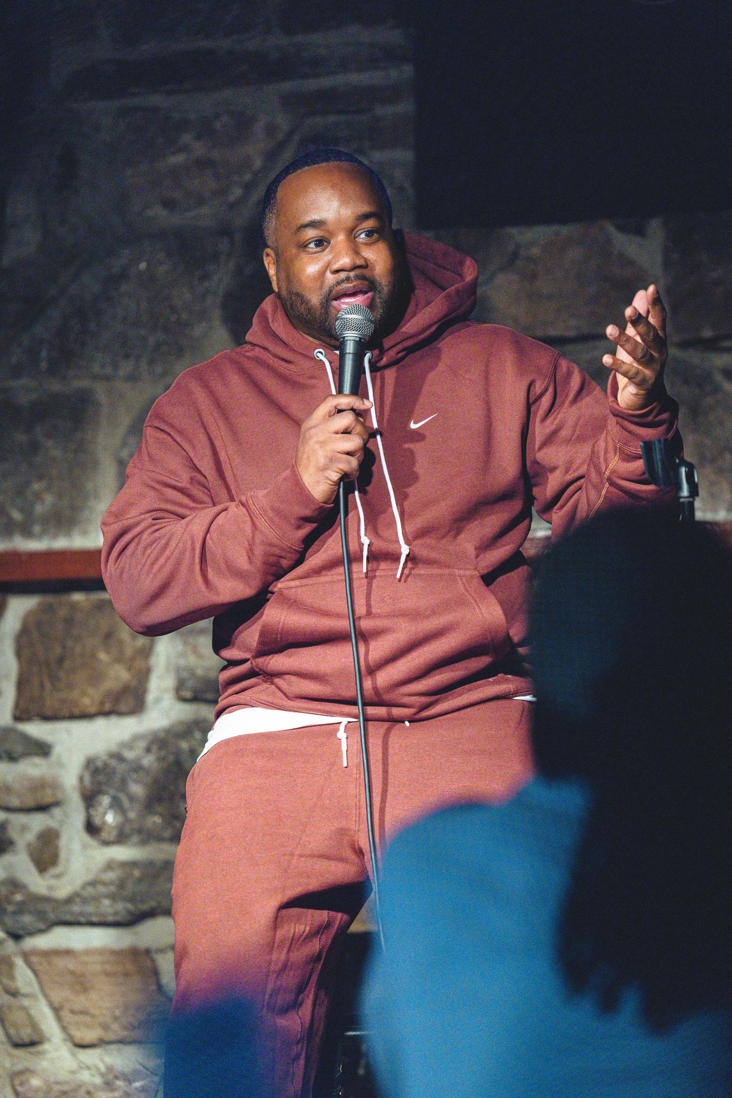 Comedian performs on stage wearing a burgundy Nike hoodie and sweatpants while gesturing with microphone in hand.