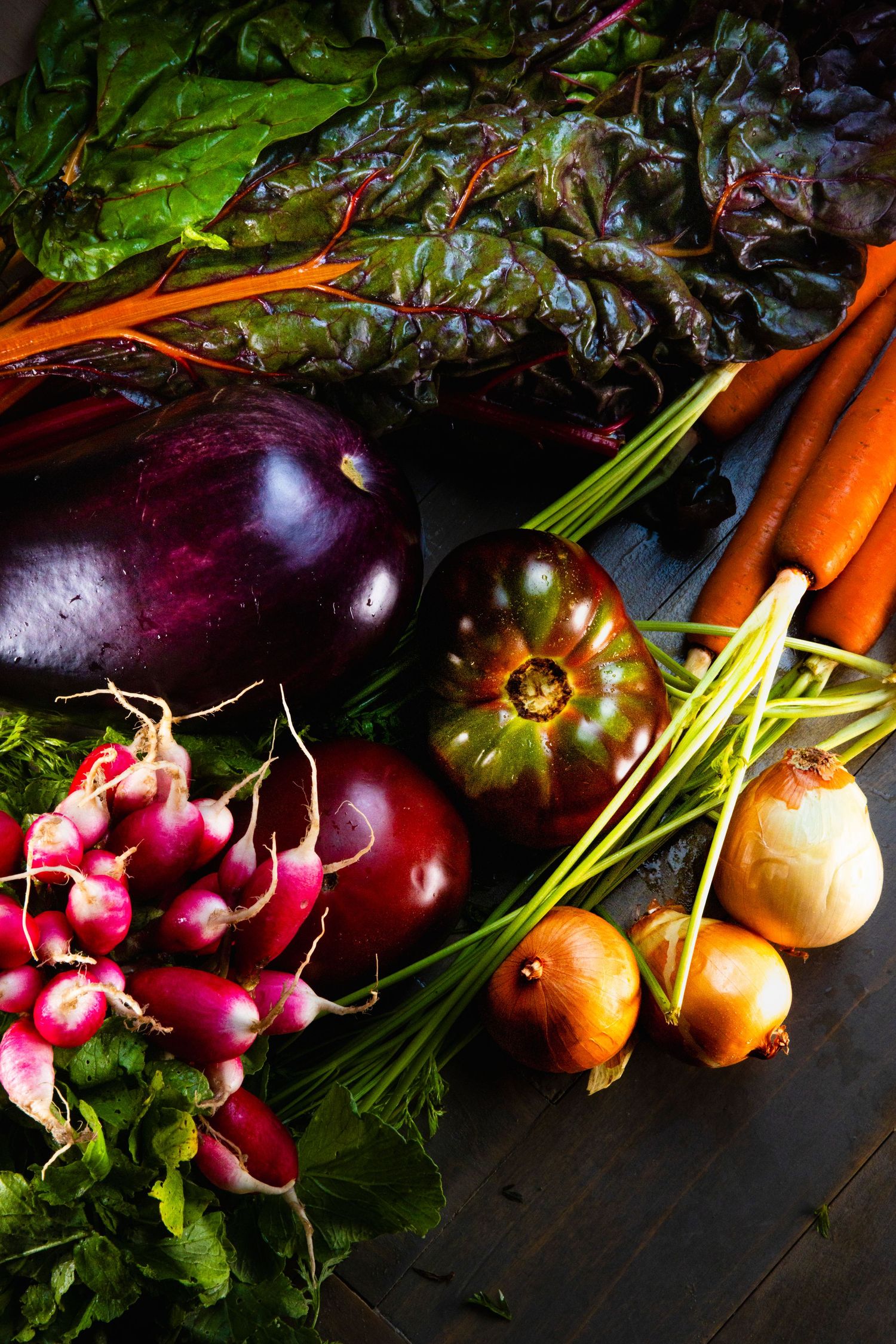 Colorful harvest of fresh vegetables including eggplant, carrots, radishes, and tomatoes on dark wooden surface.