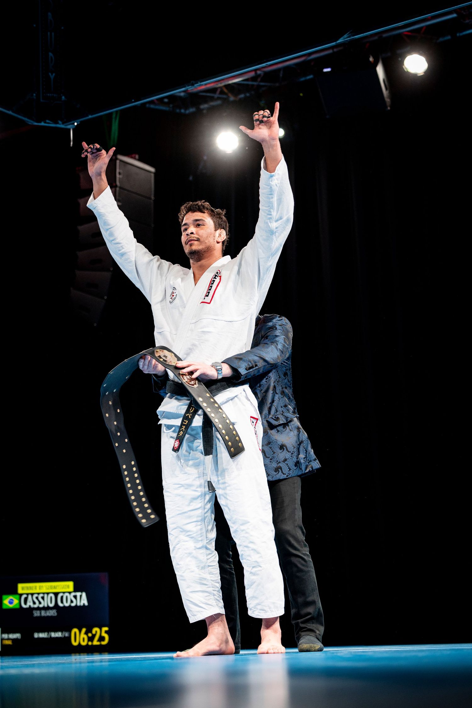 A martial artist in a white gi celebrates with raised arms after a competition victory on a dark stage.