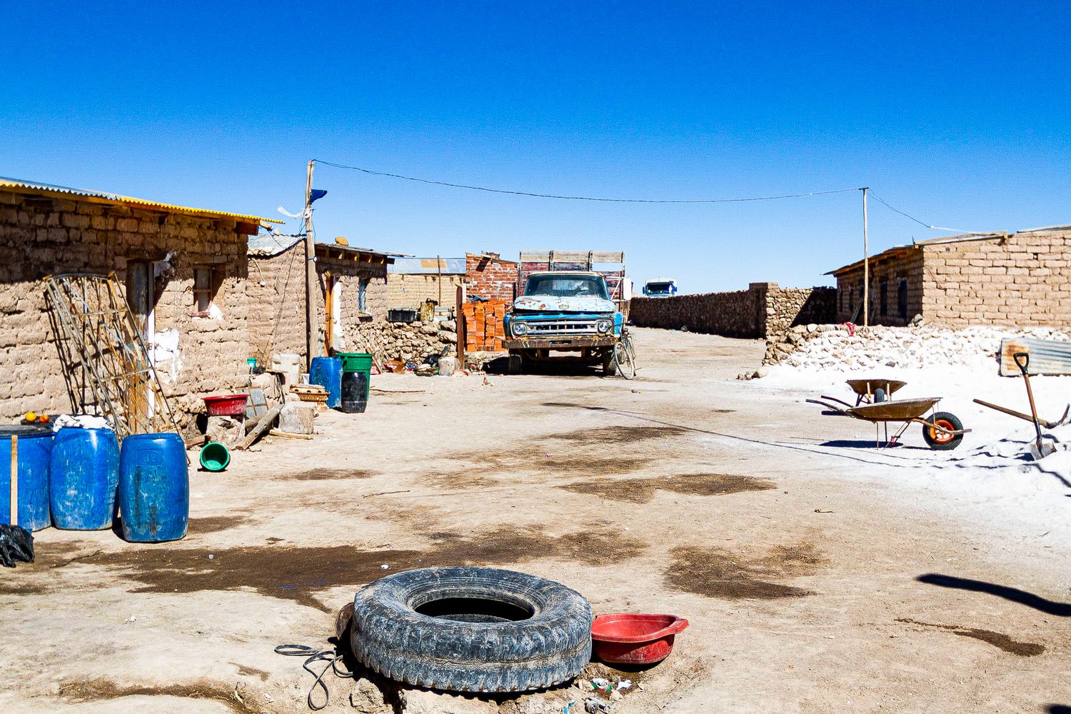 Rural desert village with blue barrels and old tire in dusty courtyard.
