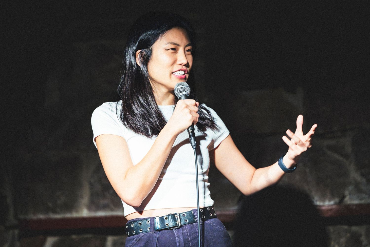 Performer in white sleeveless top and jeans gestures while speaking into microphone on dark stage.