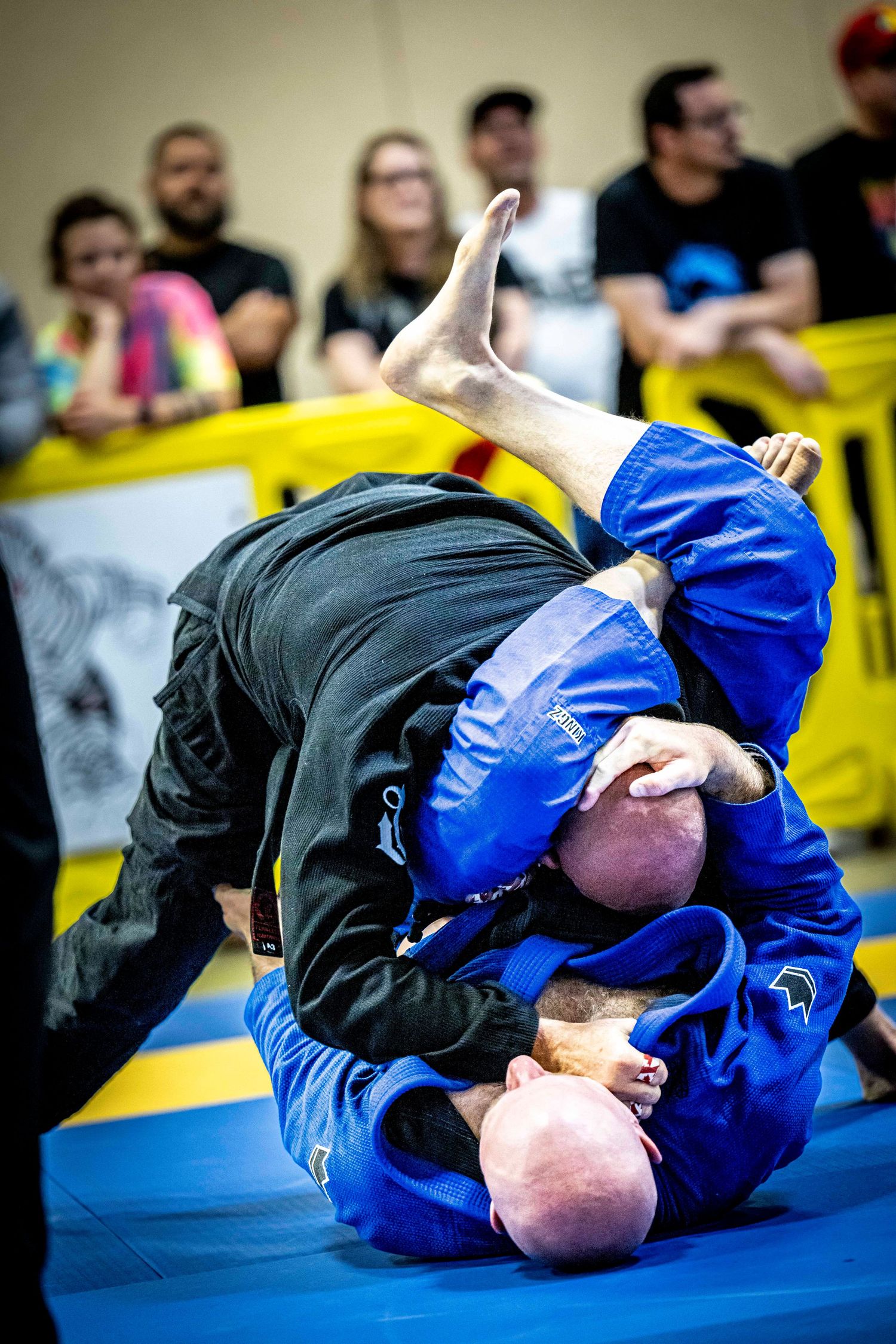Two BJJ practitioners in black and blue gis grappling during competition match.