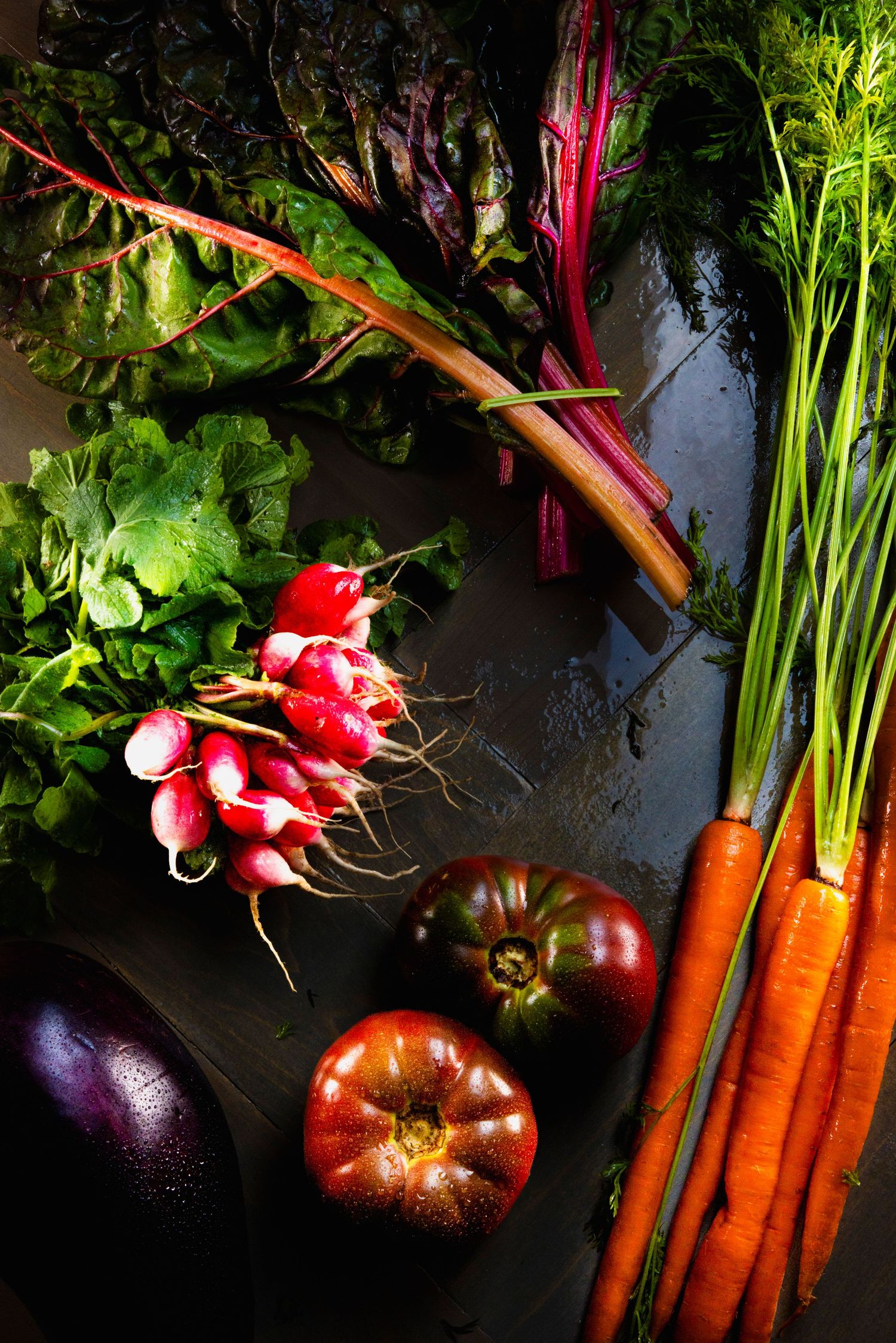 Fresh garden vegetables including radishes, tomatoes, carrots and Swiss chard arranged on dark surface in moody lighting.