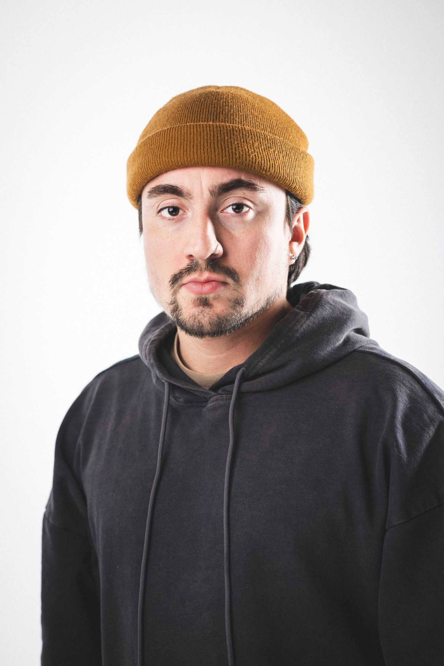 Person in black hoodie and mustard beanie looking contemplatively against white background.