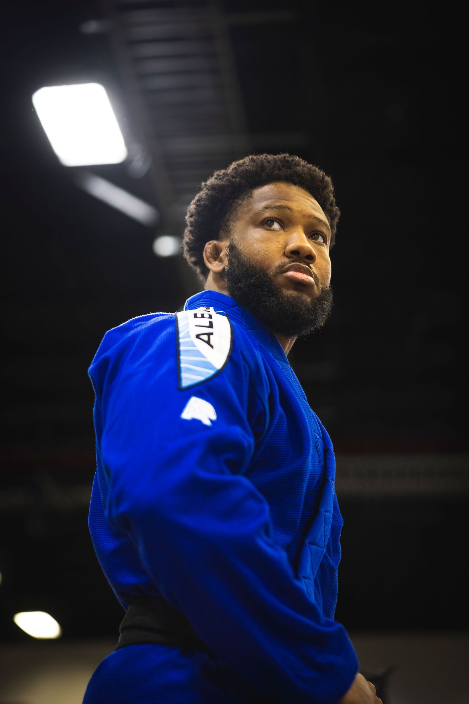 A martial artist in a blue gi stands poised during a competition under dramatic lighting.
