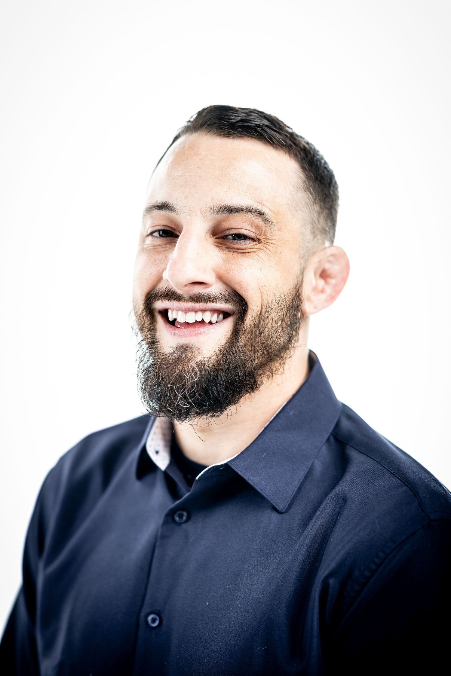 Professional studio portrait of a smiling person with dark beard wearing a navy button-down shirt against white background.