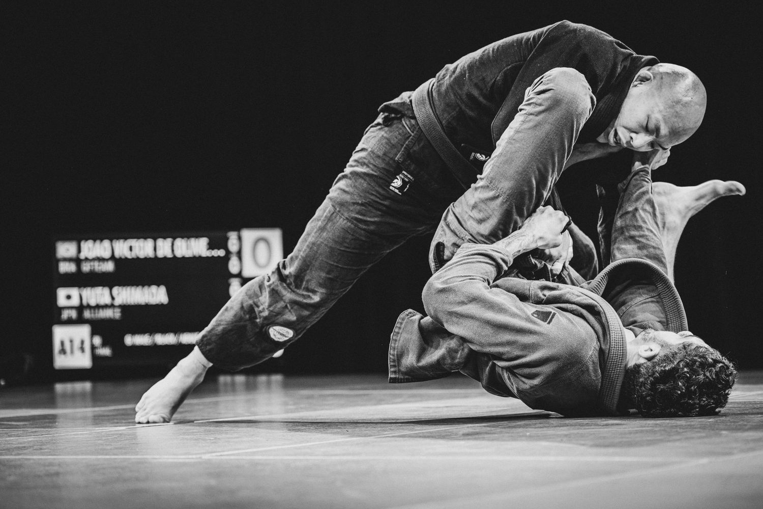 Two Brazilian Jiu-Jitsu practitioners engage in a ground grappling match, captured in dramatic black and white photography.