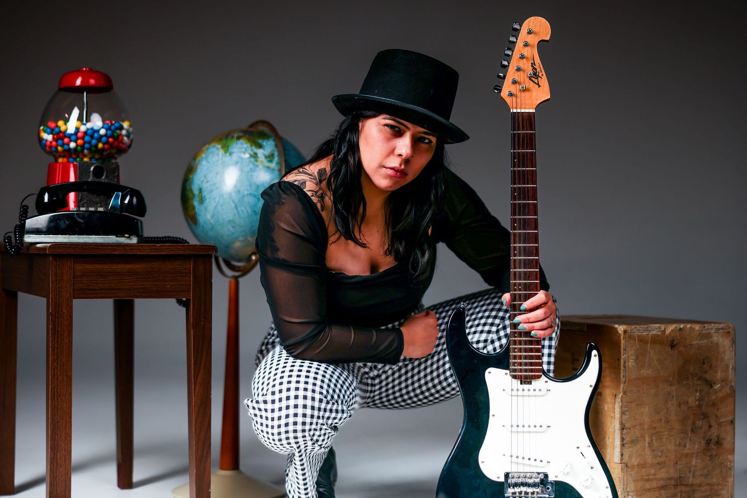 A musician poses with an electric Stratocaster guitar alongside a globe and vintage decor against a dark background.