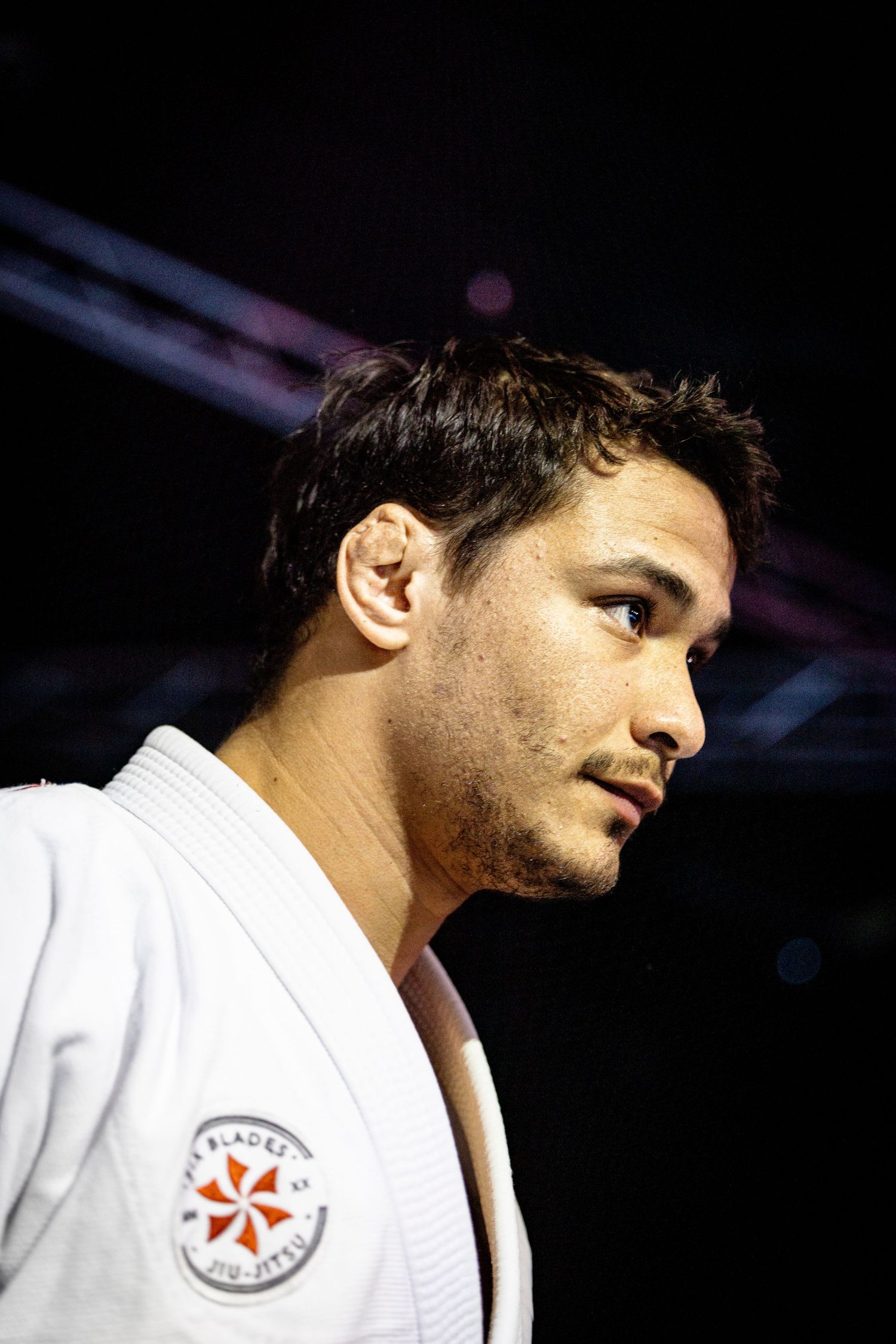 A martial artist in white gi with tournament patch stands in dramatic side profile lighting.