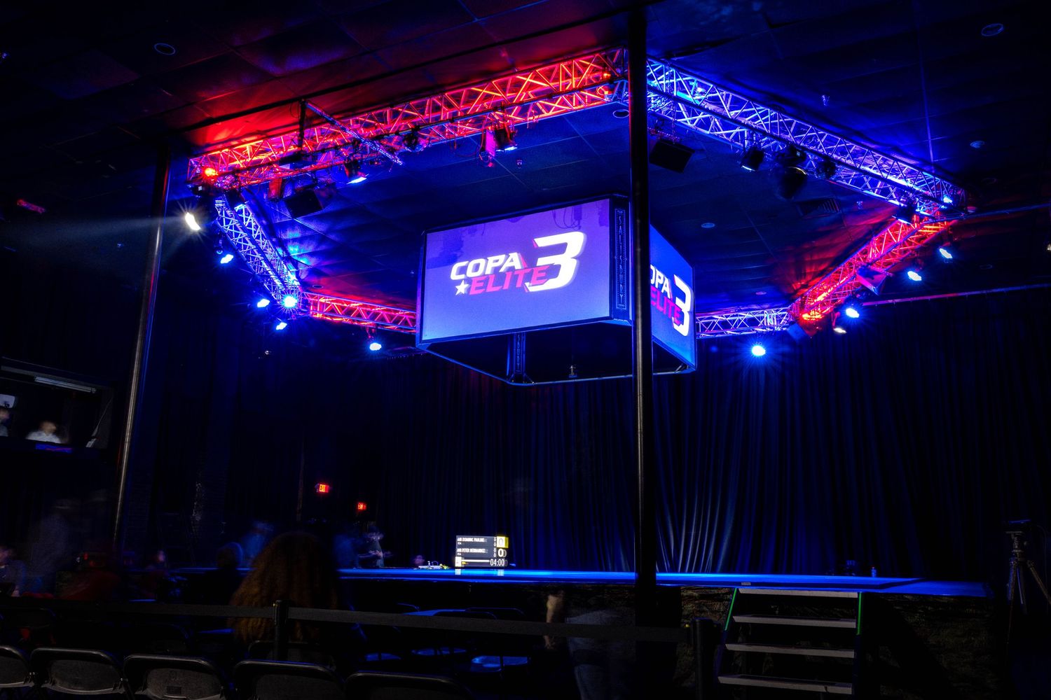 A dark stage setup with red and blue lighting featuring a digital display screen suspended from overhead truss rigging.