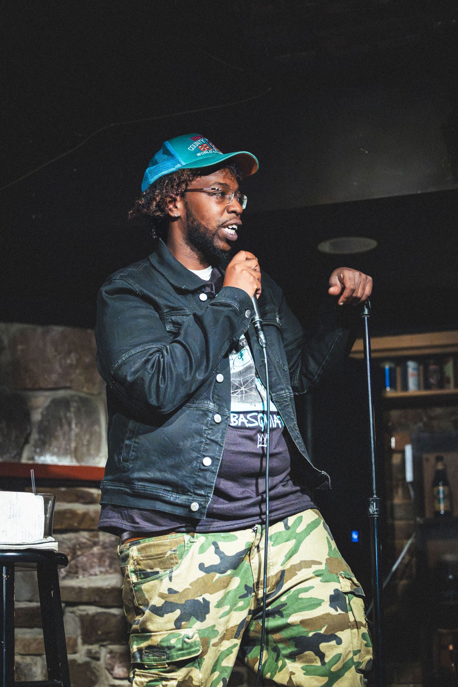 Performer in blue baseball cap and denim jacket holds microphone while wearing camo pants on dimly lit stage.
