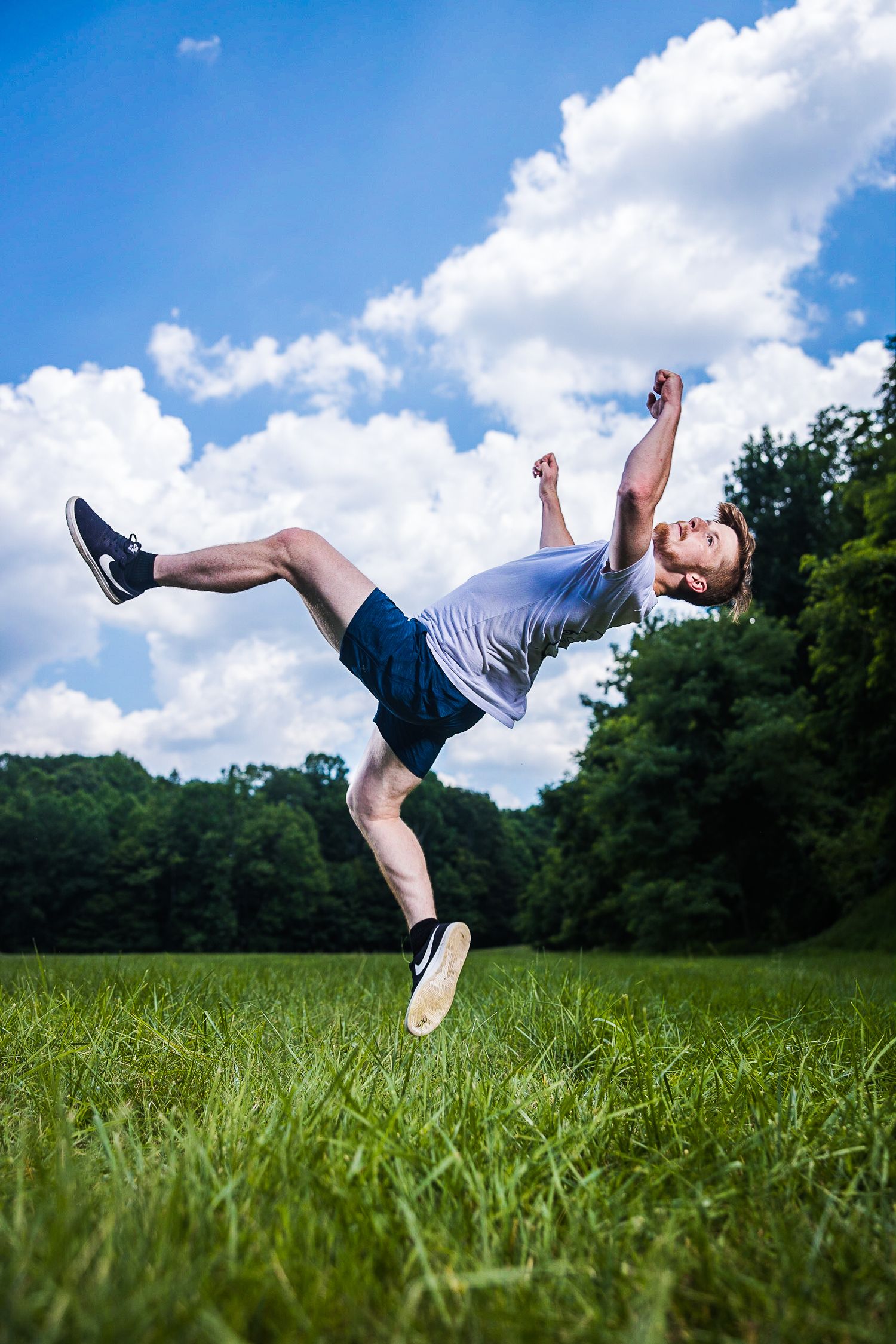 A person performs a dynamic falling or floating pose in a grassy field against a blue summer sky with white clouds.