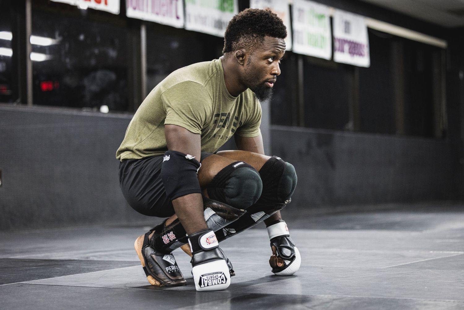 Athletic person wearing a gold shirt and dark pants crouches on a skateboard in an indoor facility.