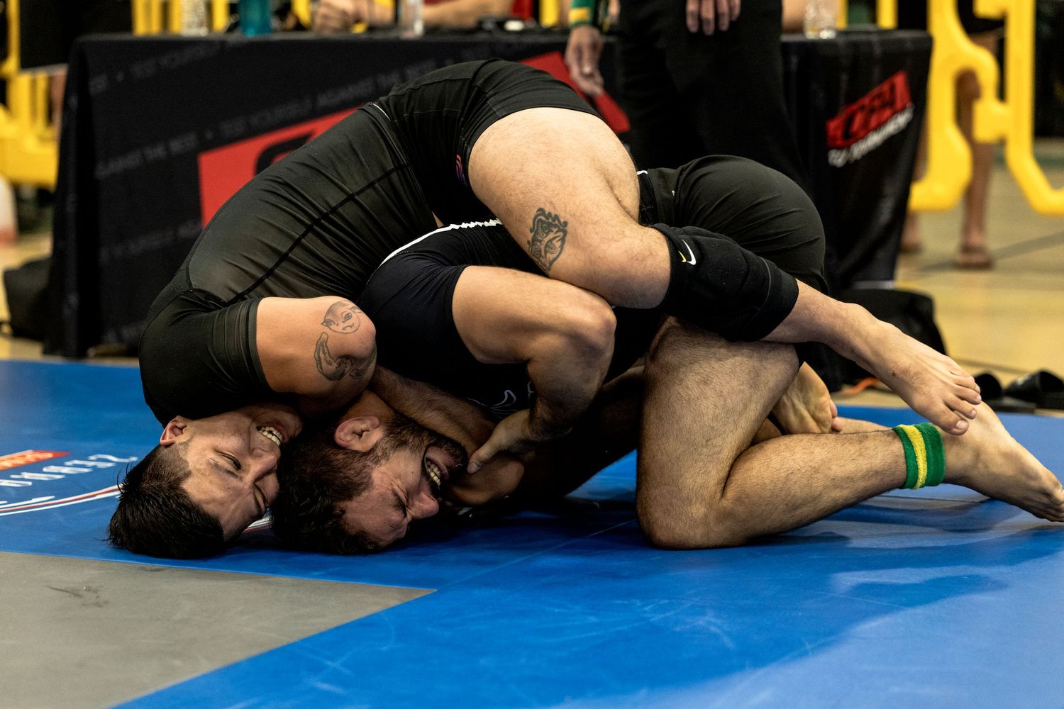 Grapplers in black uniforms compete intensely on blue competition mats during a martial arts match.