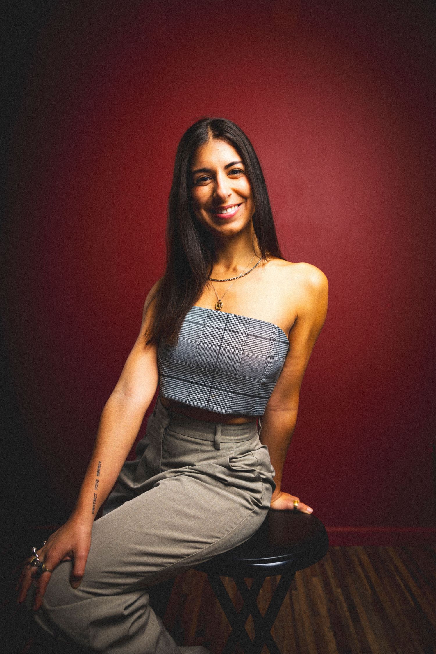 Woman in gray outfit sits casually on stool with red backdrop.