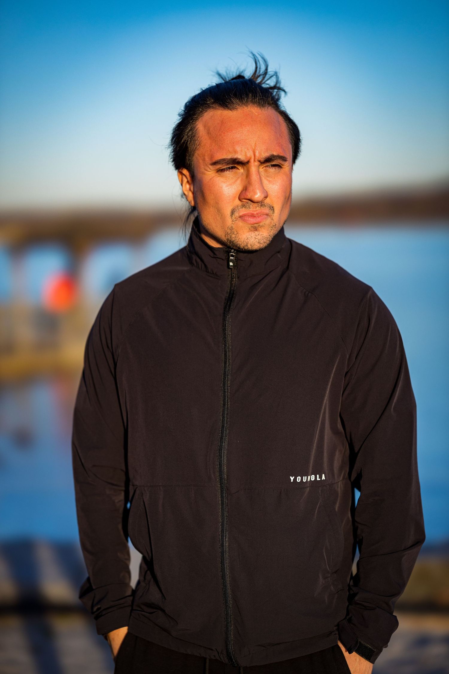 Person wearing a black zip-up jacket poses outdoors against a blurry blue background during golden hour.