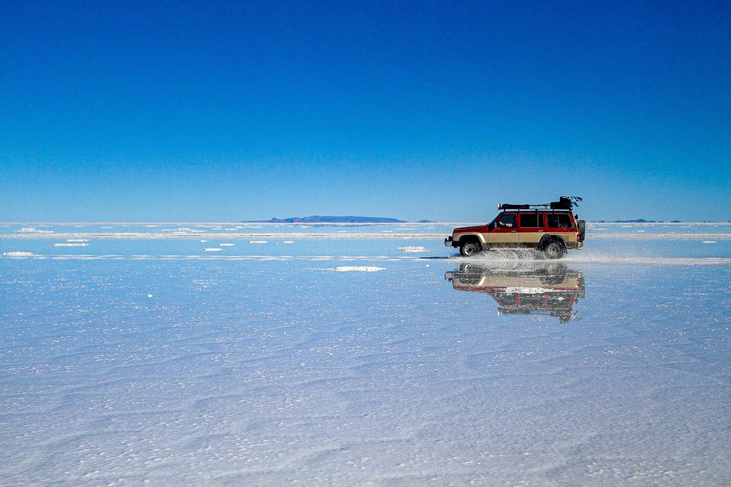 Car driving across Salar de Uyuni salt flats with perfect reflection in water.