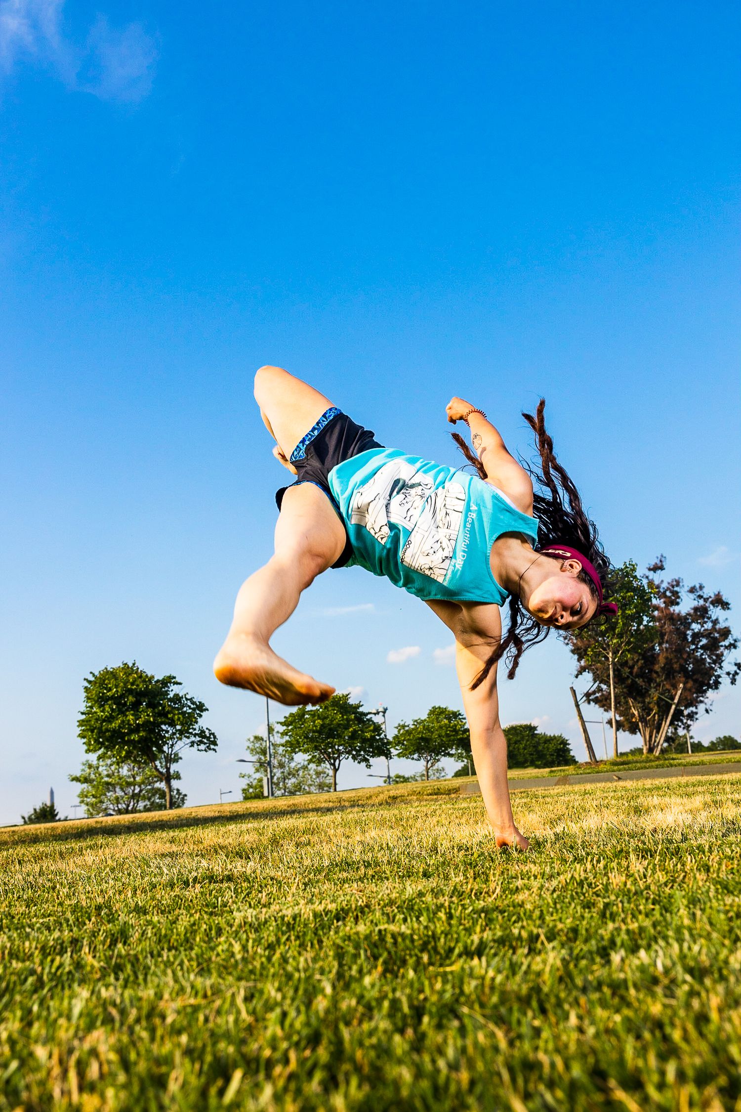 Athletic person performs an energetic breakdancing freeze move on grassy field against bright blue summer sky.