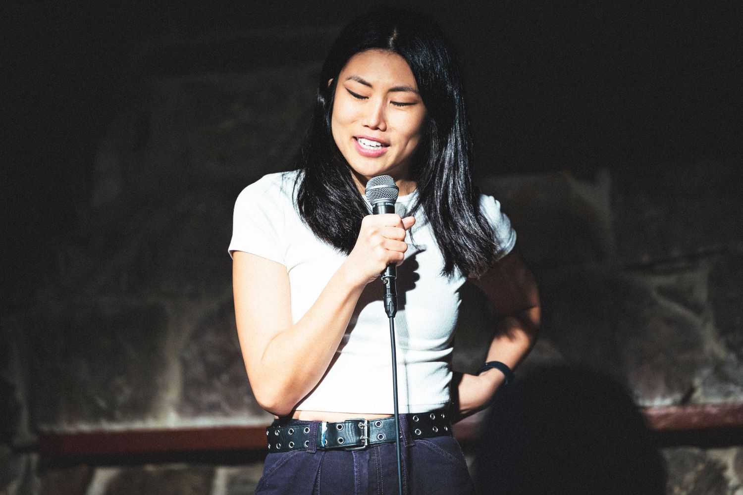 A performer in a white t-shirt and jeans sings into a microphone on a dimly lit stage.