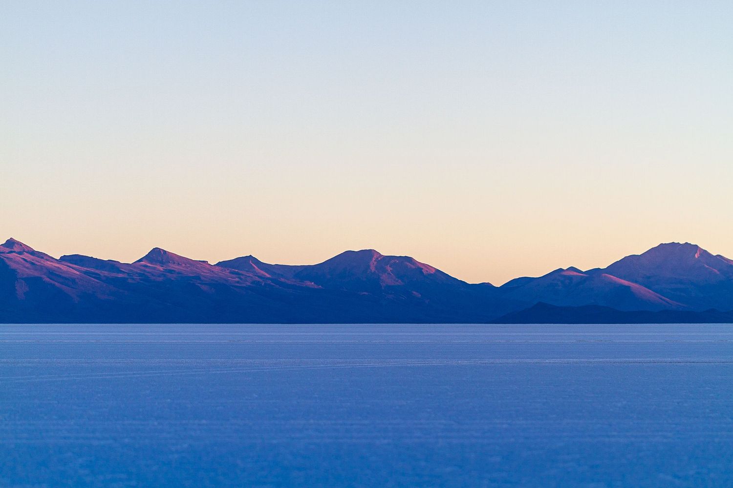 Mountain silhouettes cast deep purple hues against a pastel sunset sky over a tranquil lake at dusk.