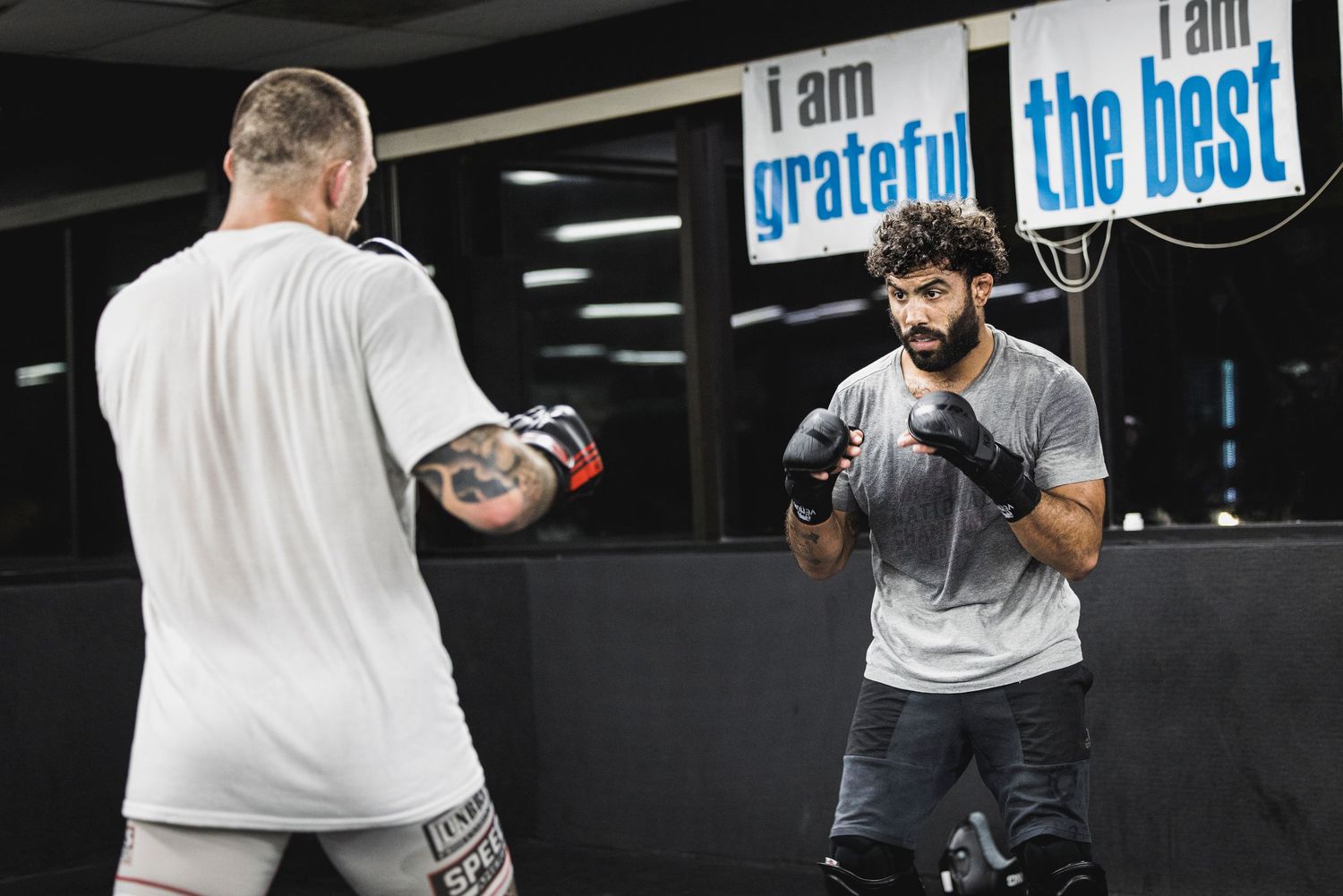 Two people practicing boxing in a gym with motivational signs reading 'I am grateful' and 'I am the best' on the wall.