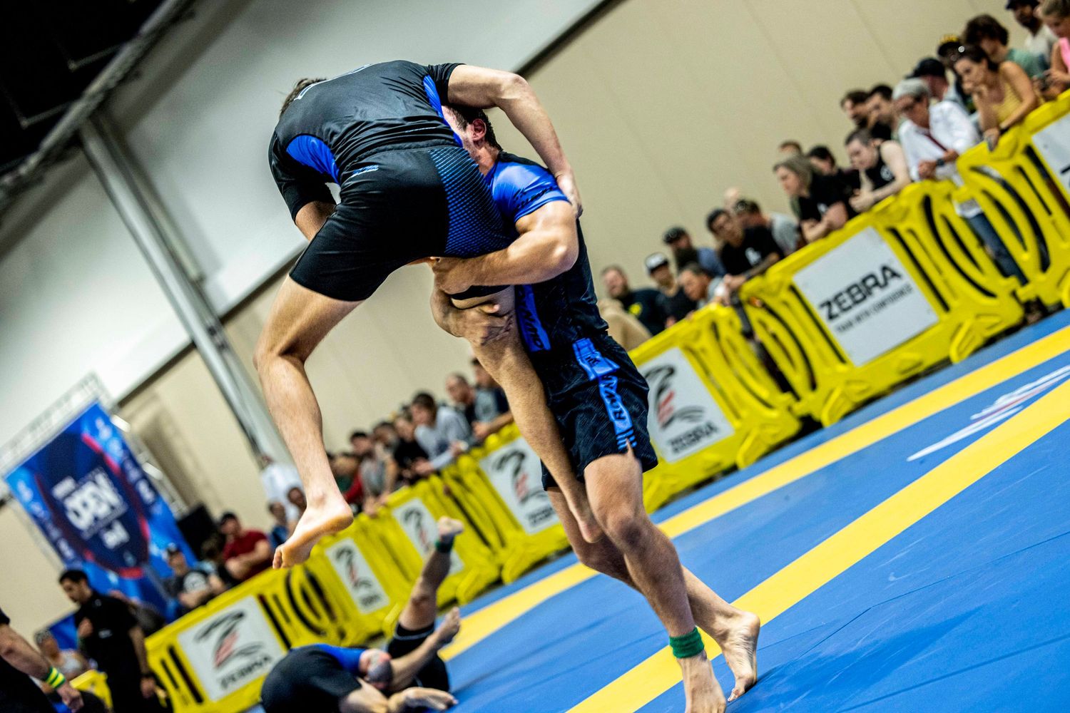 Dynamic takedown attempt during Brazilian Jiu-Jitsu competition on blue mats.