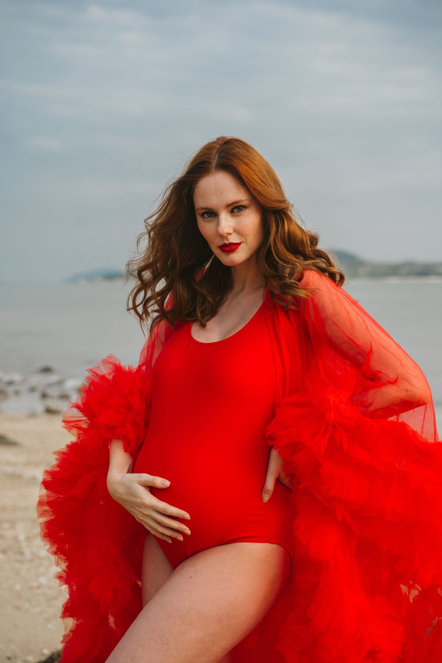 Woman in red swimsuit and flowing red tulle robe poses dramatically on a beach at sunset.