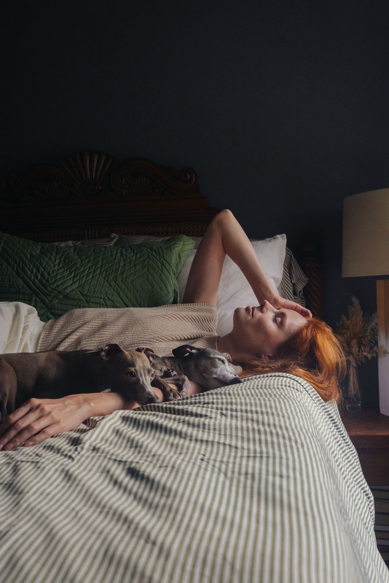 Woman and Italian Greyhound resting together on striped bedding in moody bedroom lighting.