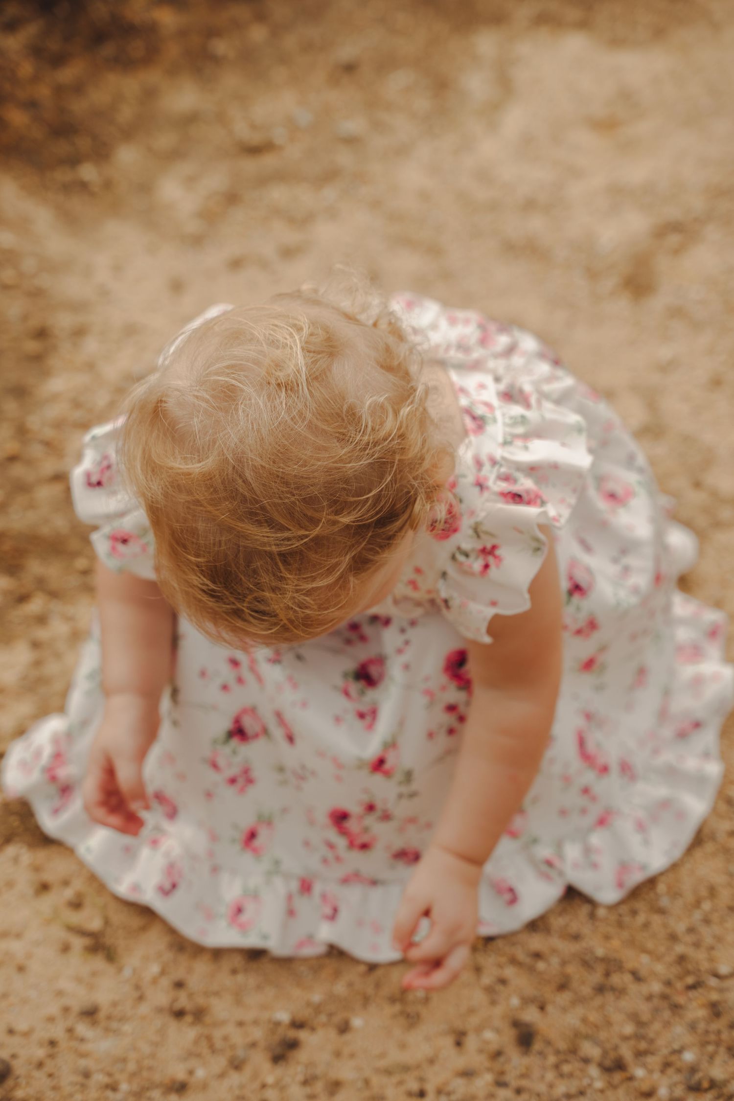 A small child in a white and pink floral dress bends down to explore something on the ground.