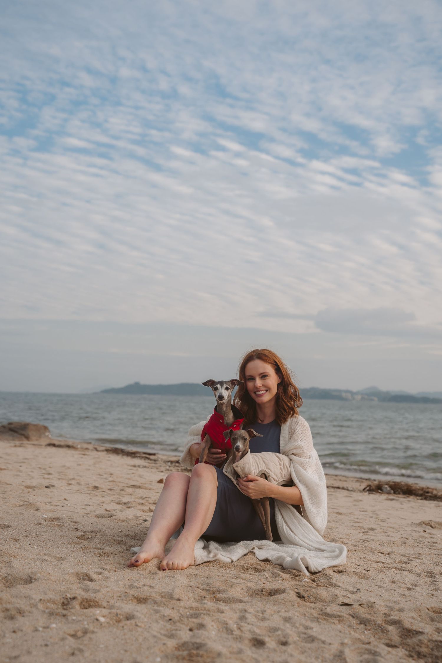Woman in white wrap sits on beach with Italian Greyhound under rippled clouds.