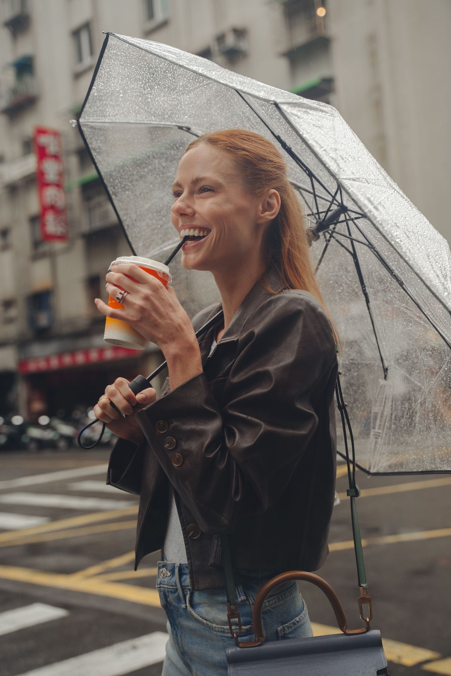 Person smiling with beverage under clear umbrella during rainy day in city.