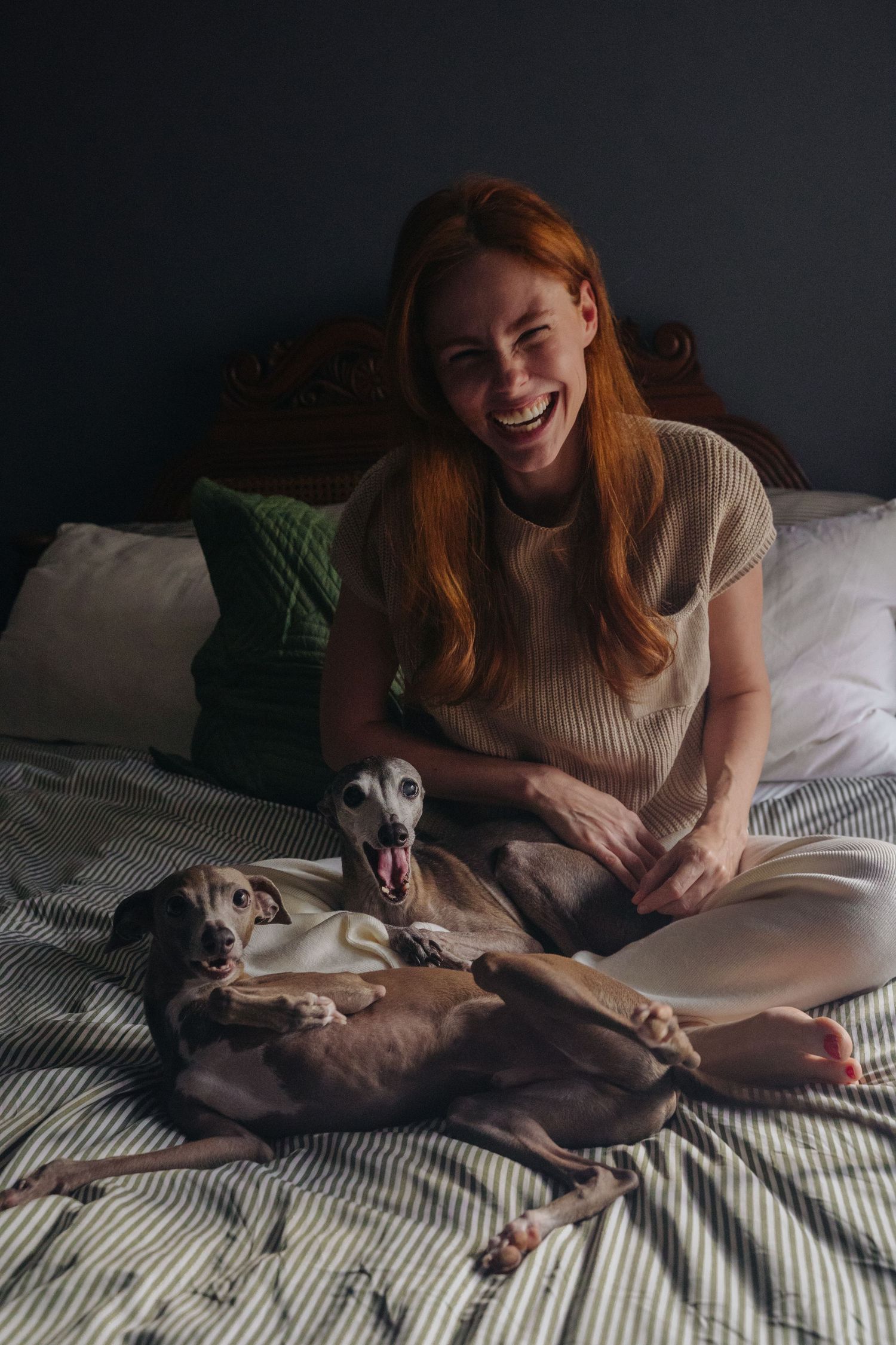 Woman sitting cross-legged on bed with relaxed Italian Greyhound in cozy bedroom setting.