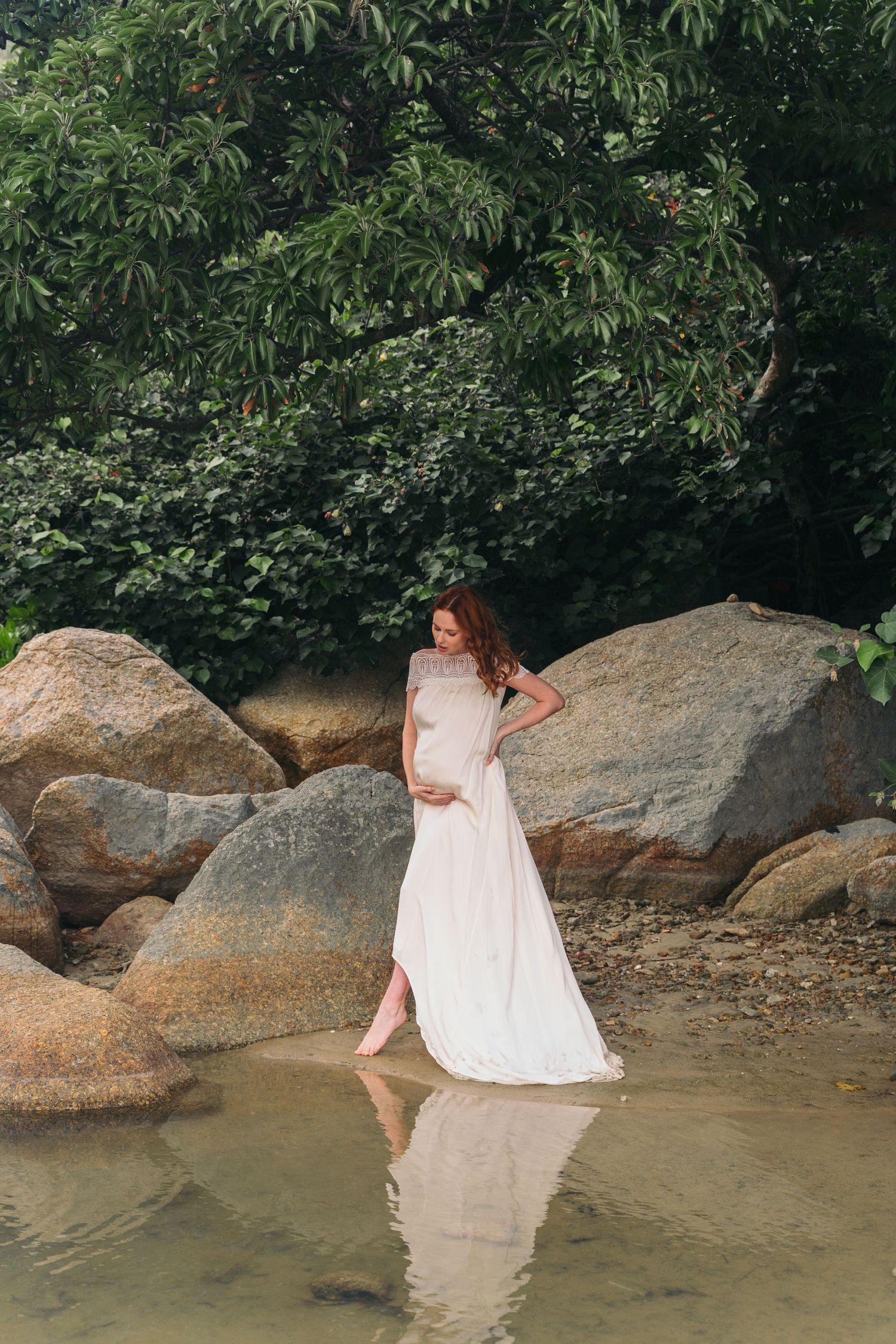 Serene maternity portrait by water with white dress and natural rock formations.