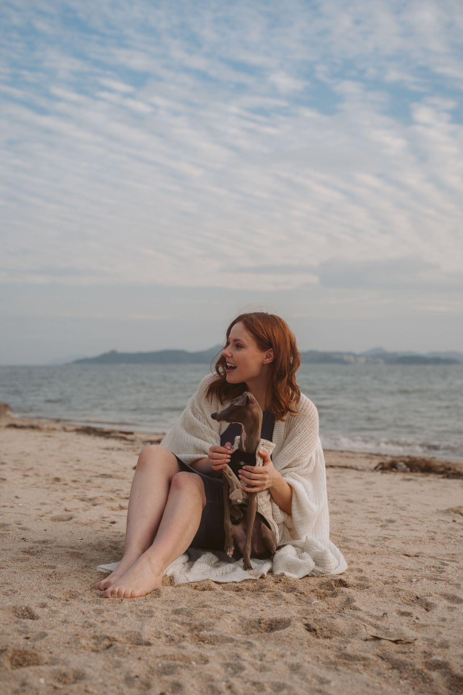 Woman cuddling with Italian Greyhound dog while relaxing on seaside beach.