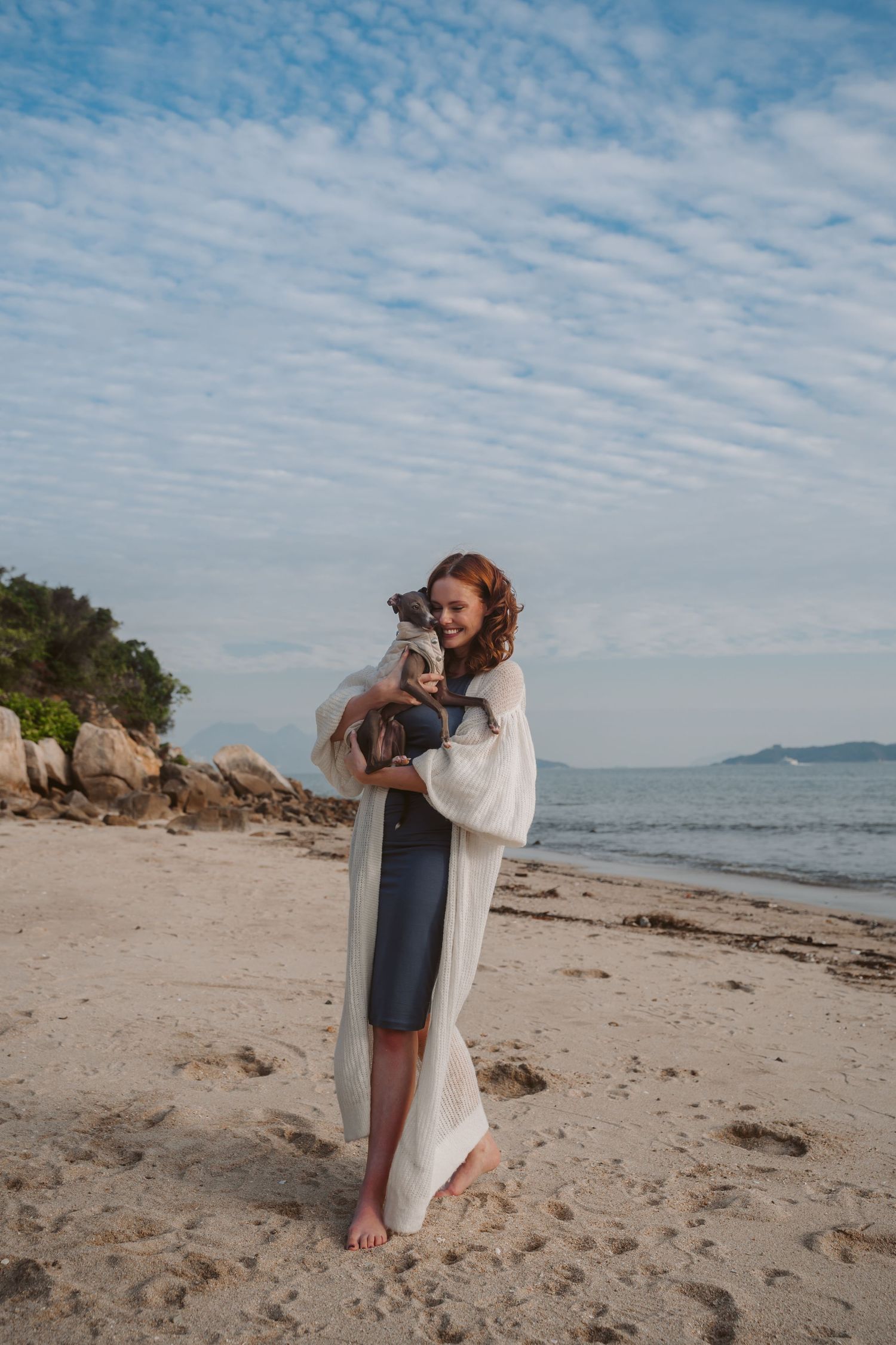 Woman in white cardigan holds Italian Greyhound on rocky beach under cloudy sky.