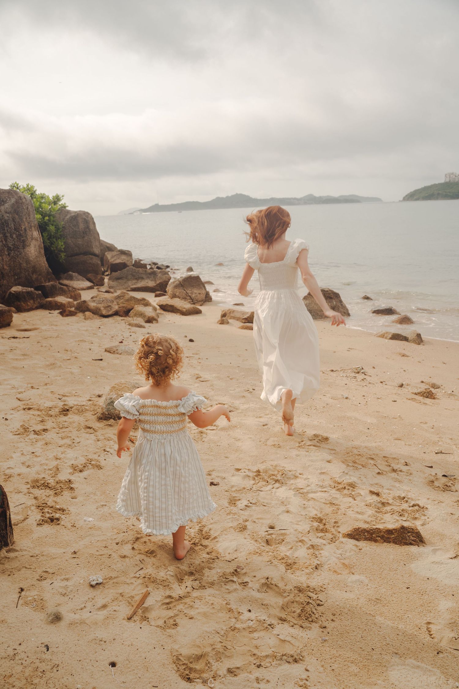 Two figures in light dresses dance joyfully along a scenic beach with rocky outcrops and distant islands.