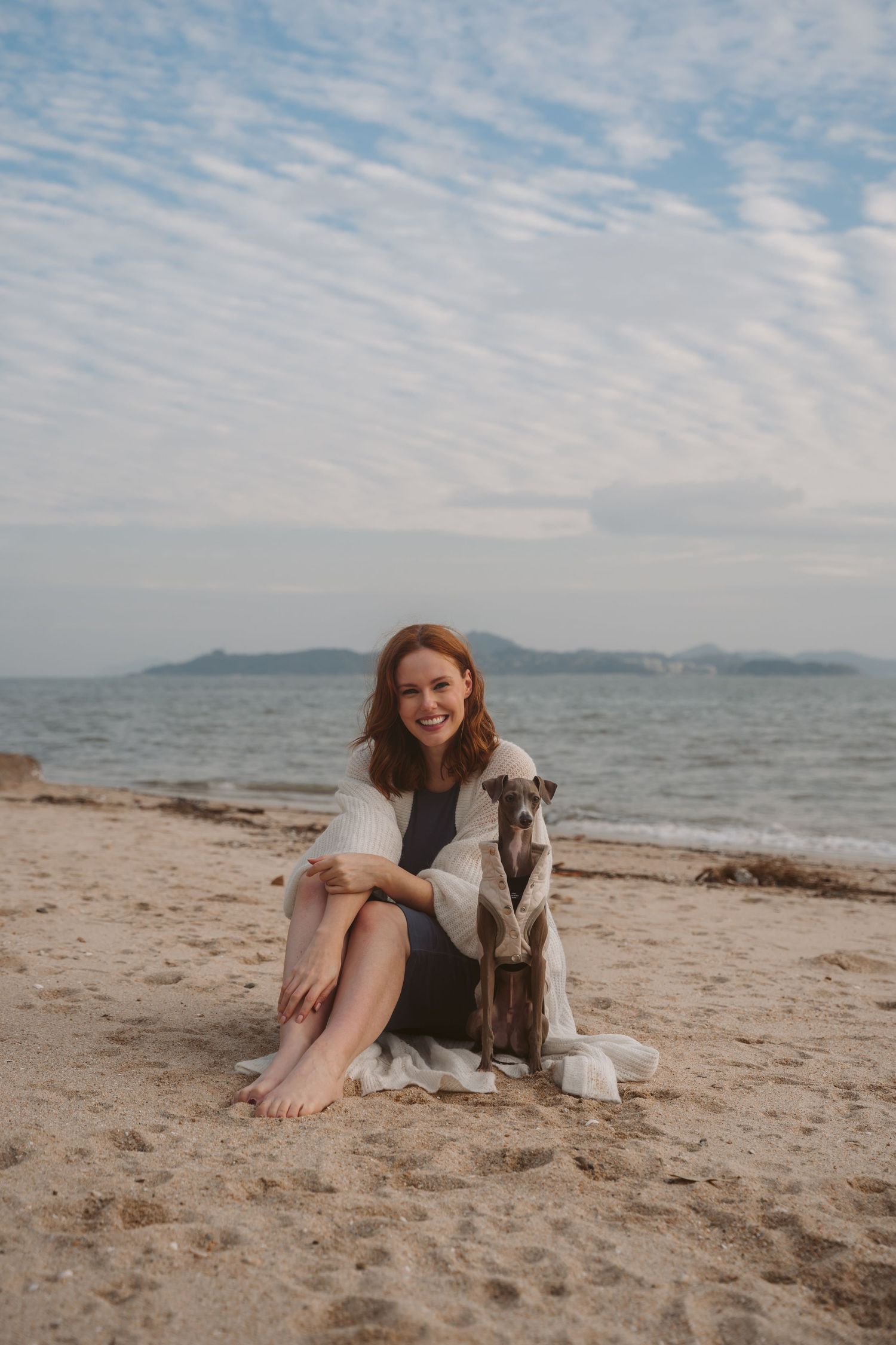 Woman in white cardigan sitting with Italian Greyhound on sandy beach with ocean view.