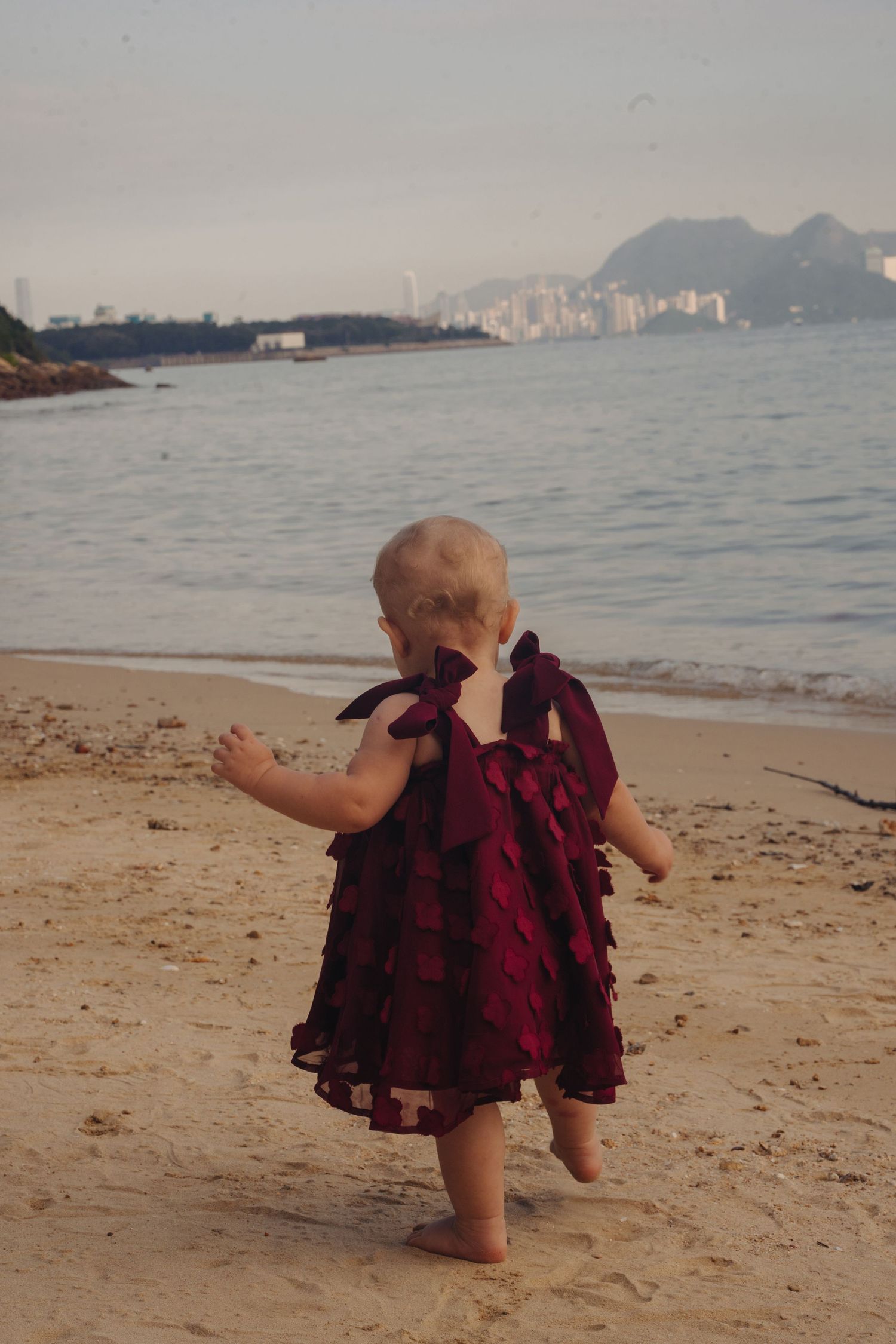 Small child in burgundy dress plays on sandy beach with city skyline visible in distance.